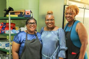 Three people in aprons smile in a colorful kitchen, standing beside shelves of supplies.