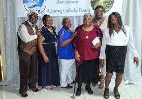 Five adults posing at a Catholic family anniversary event banner, smiling indoors