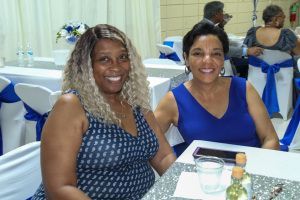 Two people smiling at a banquet table in blue dresses, with drinks and party decor nearby