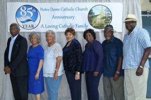 Seven adults posing in front of a Notre Dame Catholic Church 85th anniversary banner