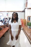 Woman in a white dress smiling in a church aisle, with pews and people seated in the background.