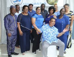 Group of people posing indoors, many in blue outfits, with one seated in front and a banner behind them.