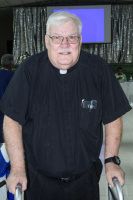 Older man in a black clerical shirt standing indoors with a screen and draped curtain behind him.