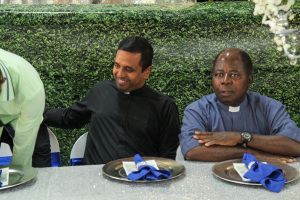 Three men seated at a table with plates and blue napkins, one smiling and touching another's shoulder.