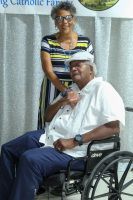 Woman stands behind seated man in wheelchair at an event, both near a white backdrop.