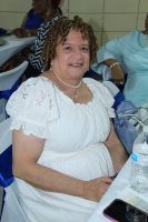 Woman in a white dress seated at a table, smiling indoors with a water bottle nearby.