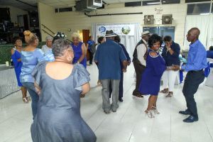 People dancing in a bright indoor hall, wearing mostly blue outfits.