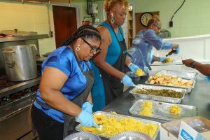 Volunteers serving food in a kitchen buffet line, wearing blue shirts and gloves