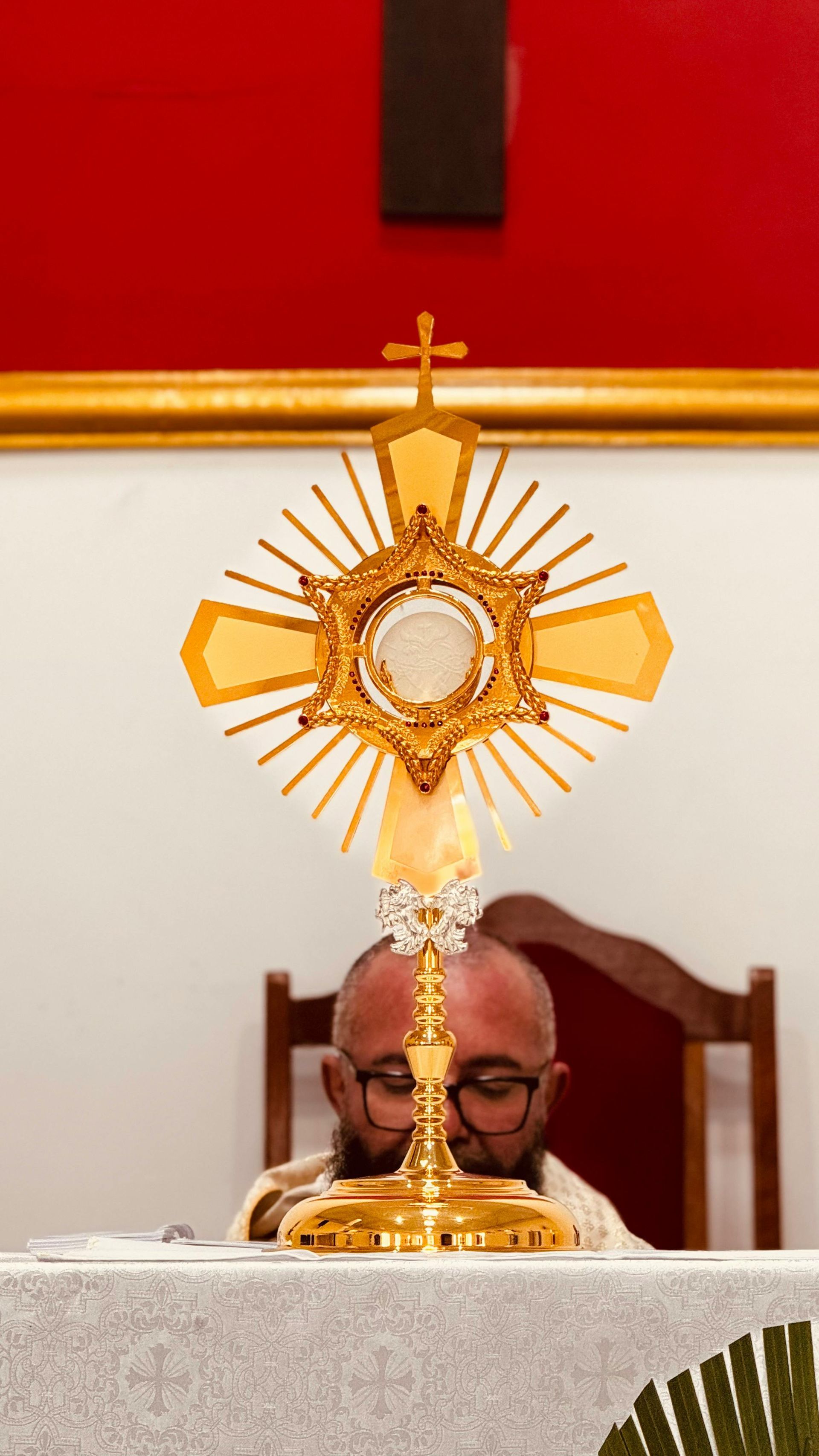 Gold monstrance on an altar in front of a priest, with a red and white church background.