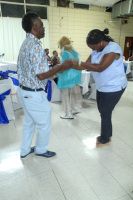 Two people dance facing each other in a bright indoor hall with chairs in the background.