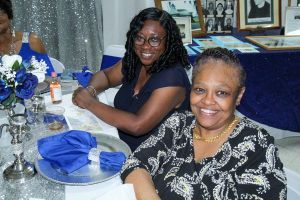 Two smiling people seated at a decorated table with blue napkins and floral centerpieces at an indoor event