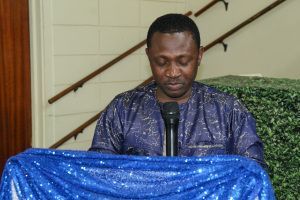 Man speaking at a podium with a microphone, draped in blue cloth, indoors near a staircase.
