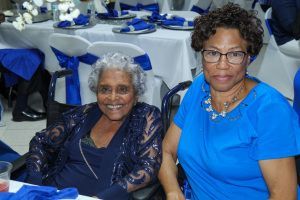 Two women seated at a banquet table, smiling in a decorated room with blue and white table settings.