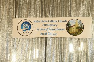 Notre Dame Catholic Church anniversary banner with blue logo and church photo on gold backdrop