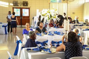 Banquet hall with guests seated at round tables dressed in white and blue, with a floral centerpiece and serving area.