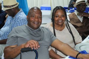 Two smiling people seated indoors, one in a gray shirt and the other in a white top, at a gathering