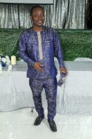 Man standing in a blue patterned suit at a decorated event backdrop with white table and floral arrangement.