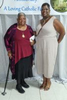 Two women posing at a Catholic event backdrop, one holding a cane, both smiling in elegant outfits.