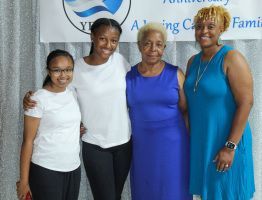 Four women standing together indoors, smiling in white, black, purple, and blue outfits.