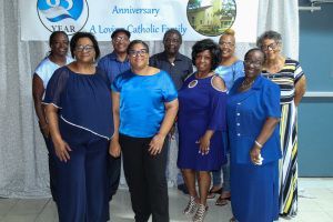 Eight women posing in blue outfits at an anniversary celebration banner.