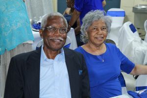 Two smiling people seated at a formal indoor event, one in a dark suit and one in a blue dress.