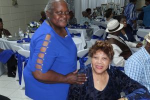 Two women at a banquet table, one standing in a blue shirt and one seated in a dark jacket, smiling