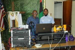 Two men stand behind a table of electronics and laptops in a room with flags and a wooden podium.