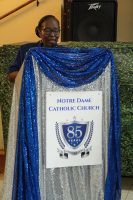 Man standing behind a blue-and-silver draped podium with a sign reading “Home Dame Catholic Church 85”