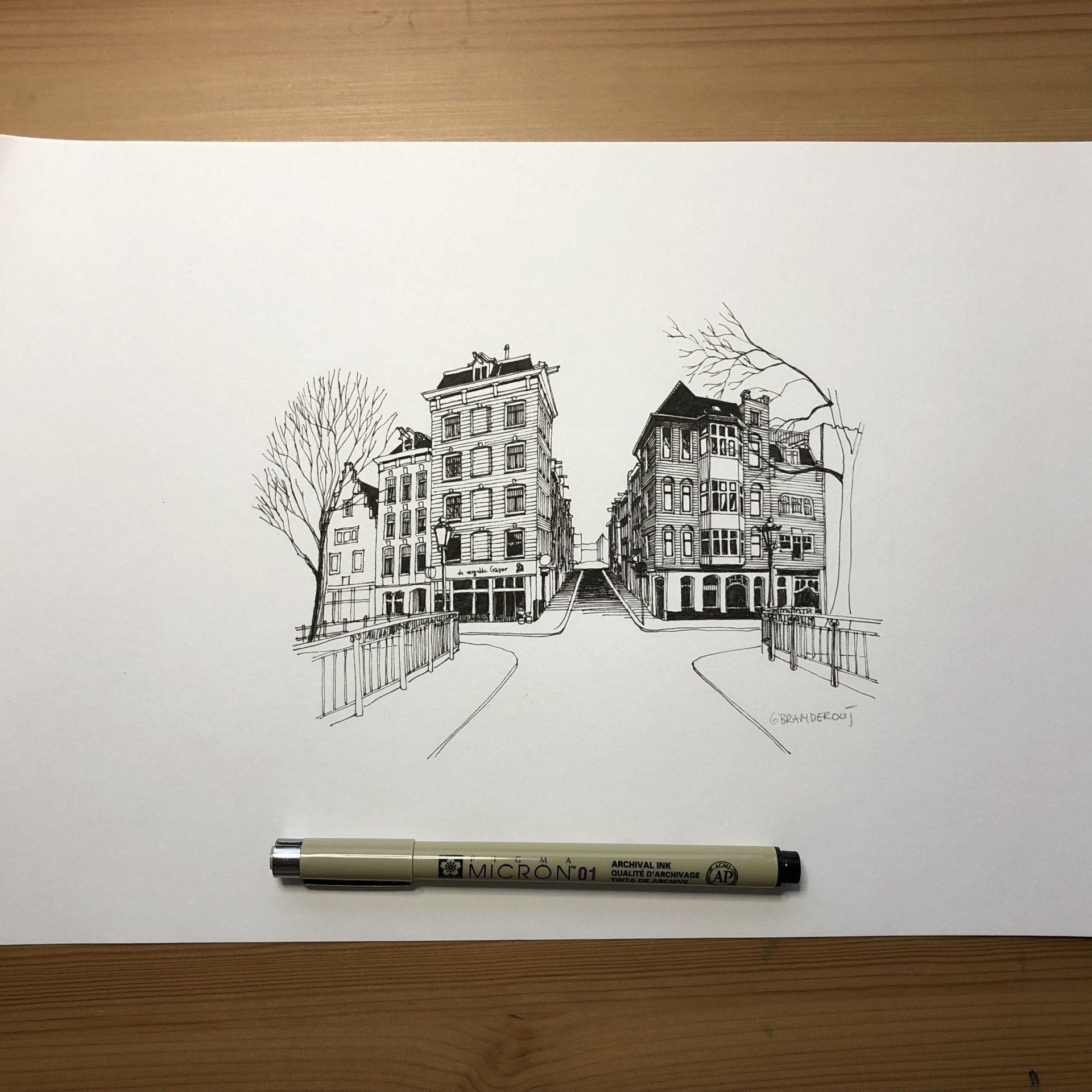 Inkttekening van twee gebouwen op wit papier, met een pen eronder op een houten tafel.