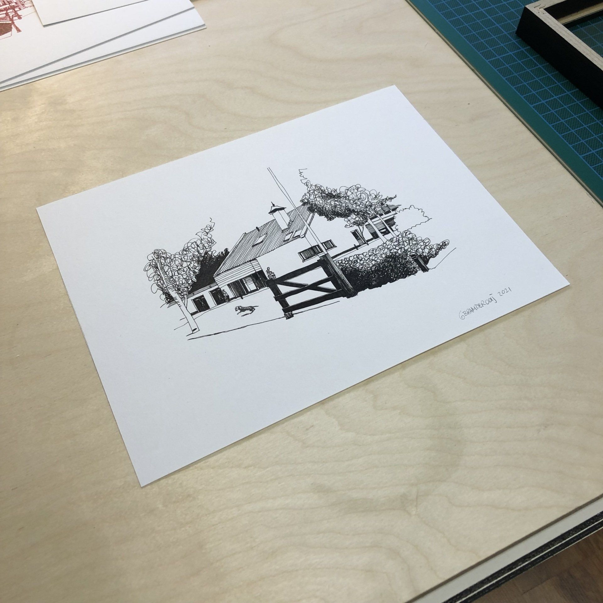 Zwart-wit schets van een huis op wit papier, op een bureau naast een snijmat.