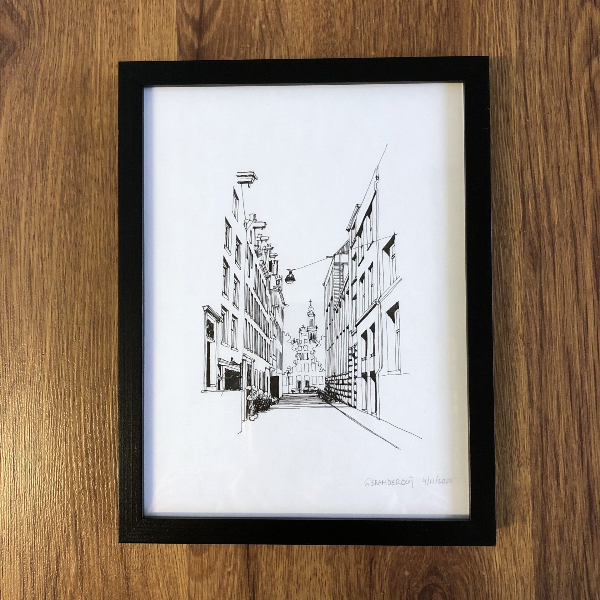 Ingelijste zwart-wit schets van een smalle stadsstraat tussen hoge gebouwen op een houten ondergrond.