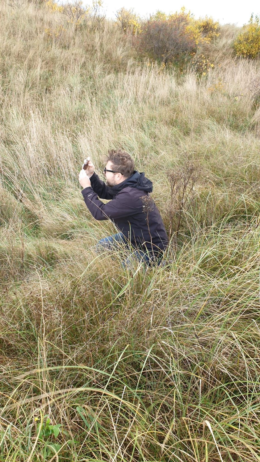 Bram de Rooij knielt in het hoge gras en houdt een telefoon omhoog om buiten een foto te maken.