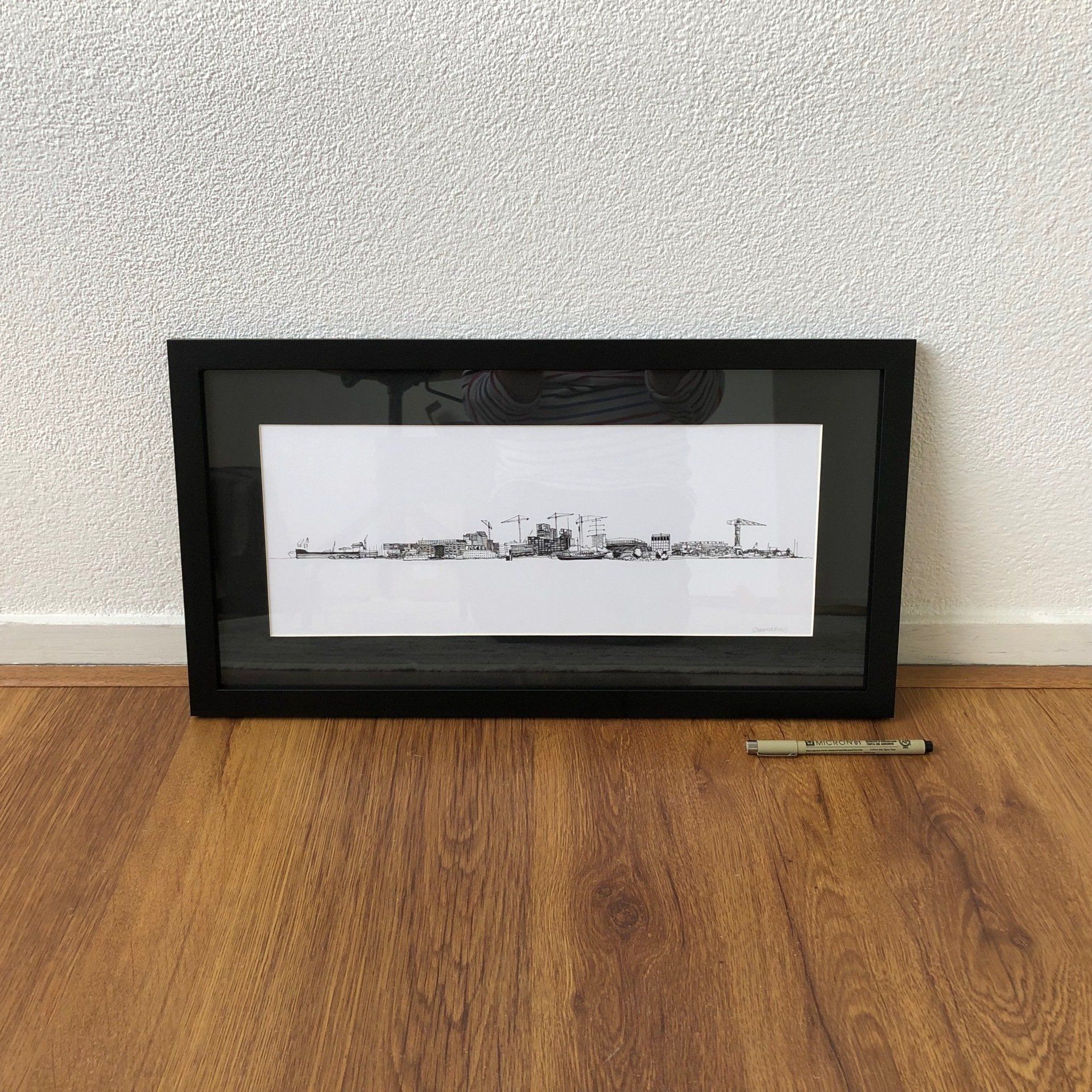 Een ingelijst zwart-wit panorama leunt tegen een muur op een houten vloer, met een potlood ernaast.