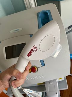 Hand holding a white laser hair removal device in front of a larger machine with a blue and white case.