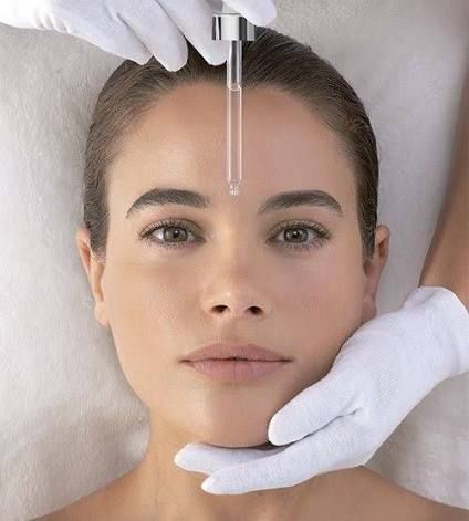 Gloved hands administering a facial skincare treatment with a dropper to a woman lying down