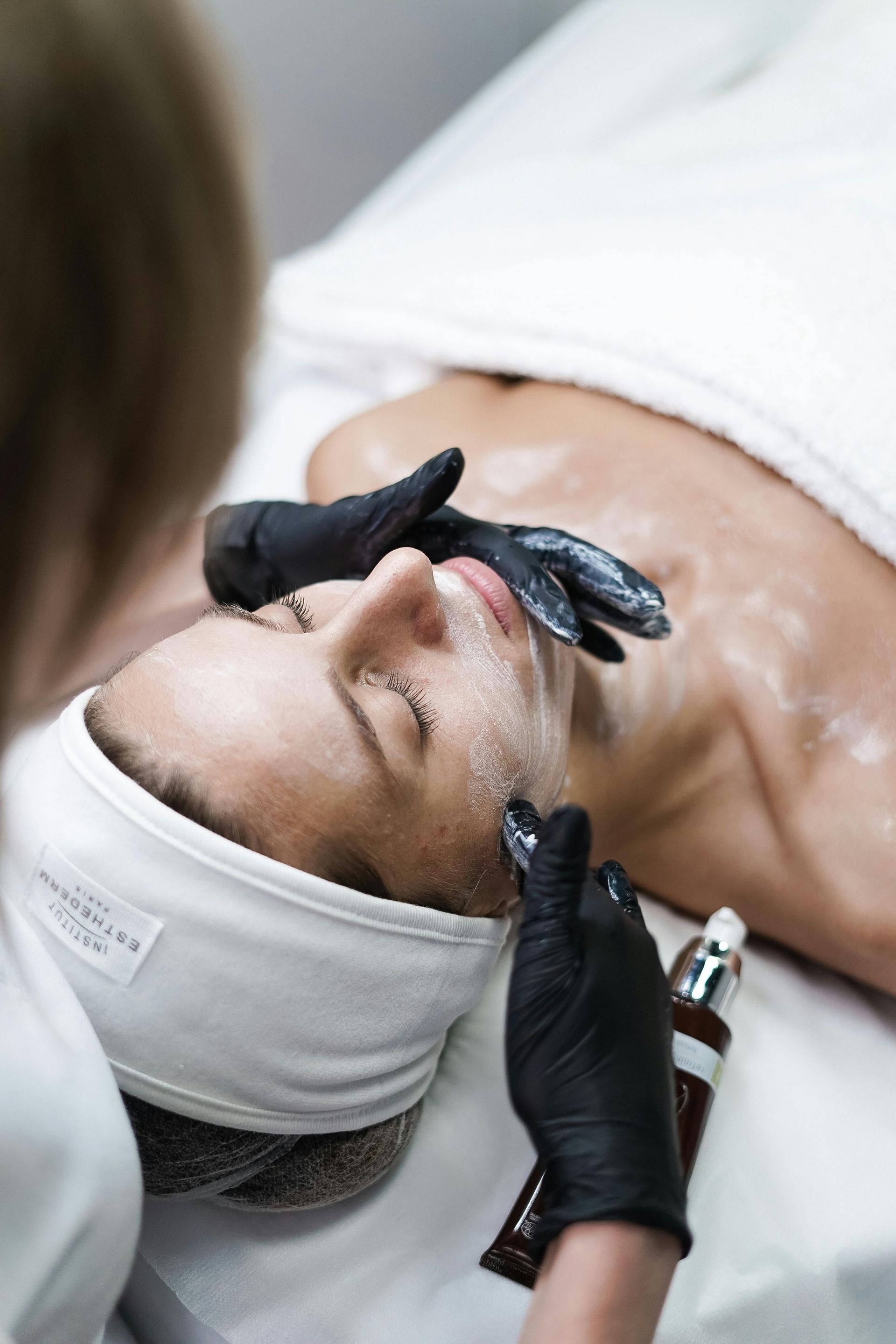 Person receiving facial treatment at a spa; esthetician applying product with gloved hands.
