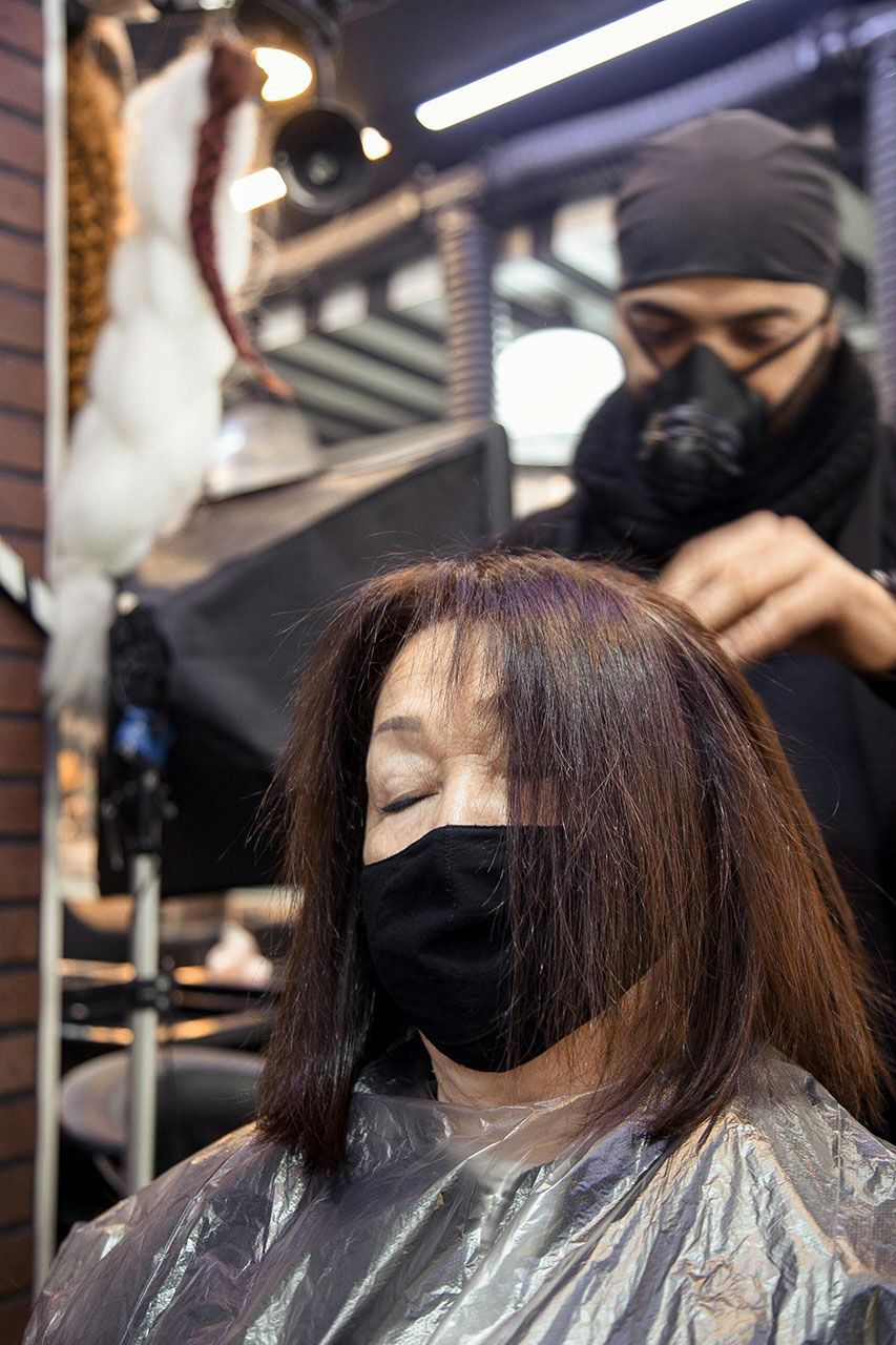 Person with mask gets hair styled by stylist wearing face mask and head covering in a salon.