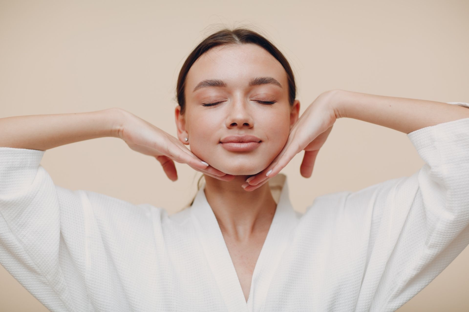 Person in white robe with eyes closed, gently massaging face against a beige background