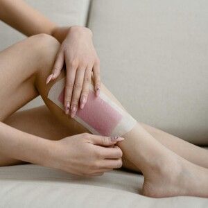 Person applying wax strip to their leg while sitting on a light-colored couch.