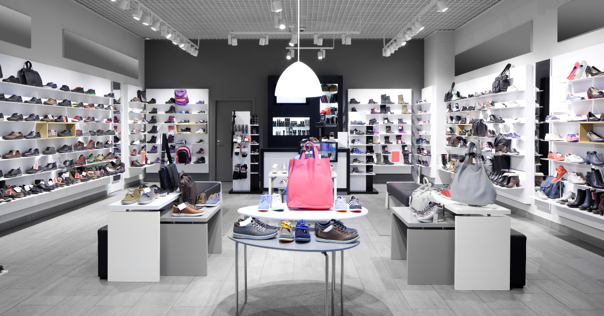 Shoe store interior with shelves displaying various shoes. A central table holds purses; neutral walls.