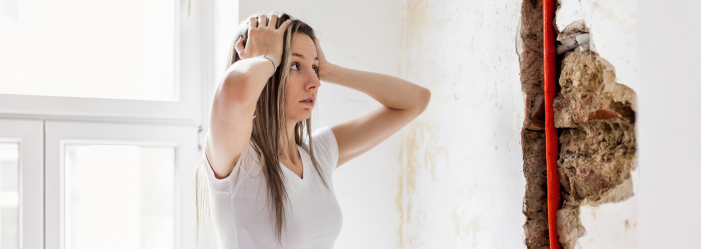 Woman looking distressed at damaged wall with exposed pipe.