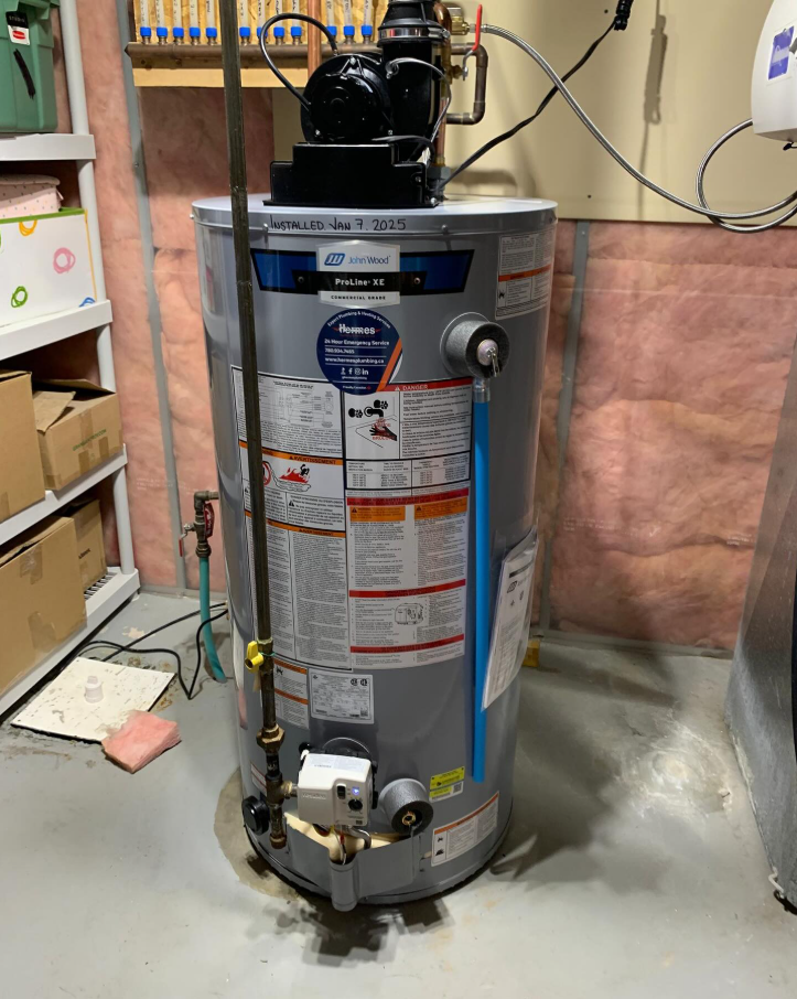 Water heater in a basement. The unit is metal, with pipes and a black top, sitting on a concrete floor.