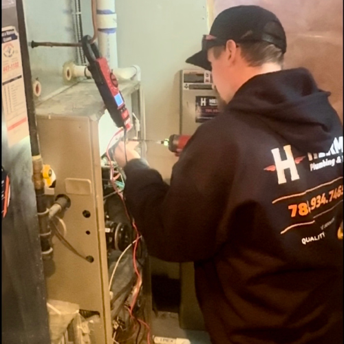 Technician in a branded hoodie uses a multimeter and power drill to repair a furnace in a residential utility room.