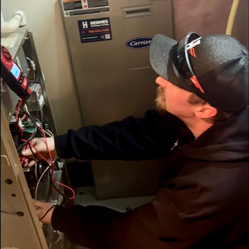 A technician in a black hoodie and cap uses a multimeter to test electrical wires inside a Carrier furnace unit.