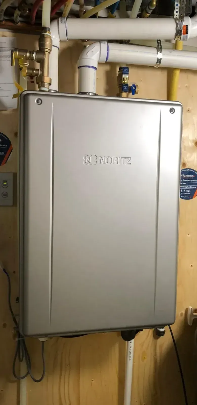 A silver water heater is hanging on a wall next to pipes.