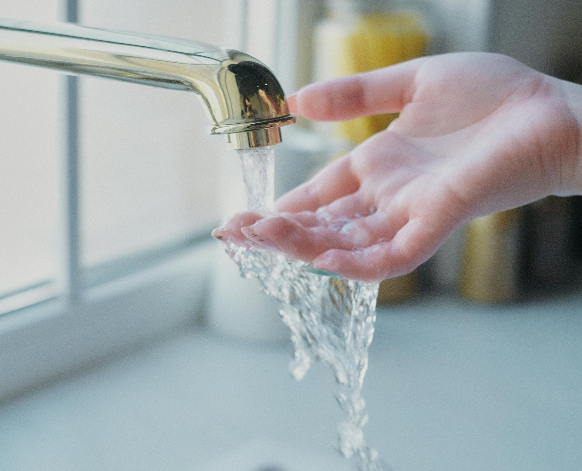 Hand under running faucet, water flowing into the palm, near a window.