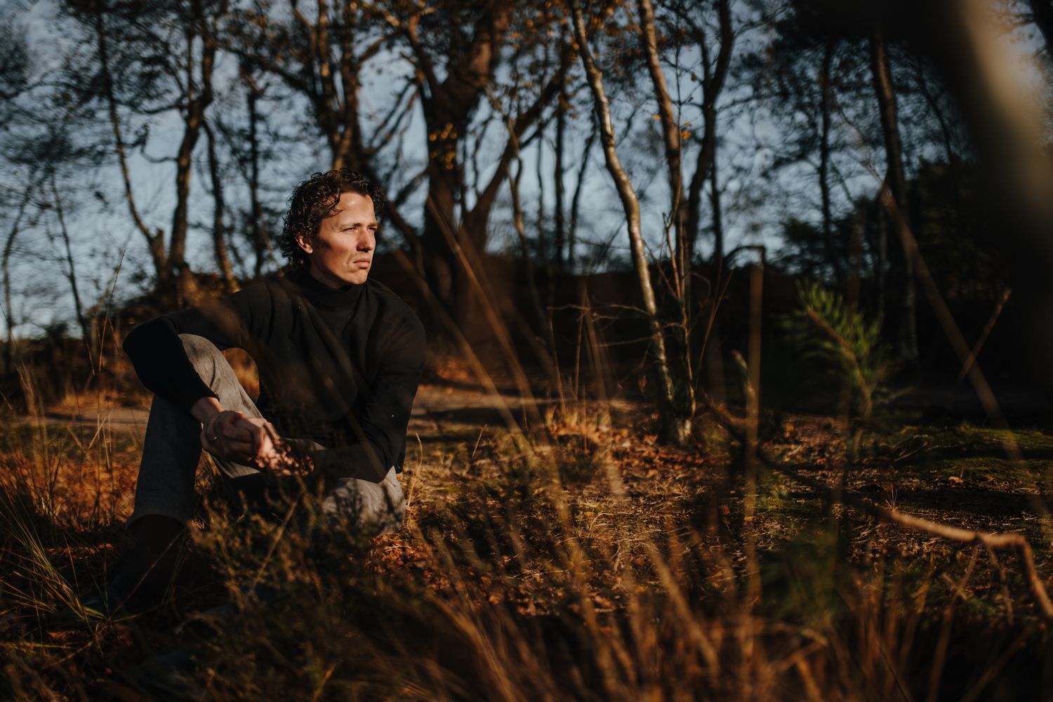 Een persoon zit in de schemering op een zonnige, grasrijke open plek in het bos en kijkt met een peinzende uitdrukking opzij.