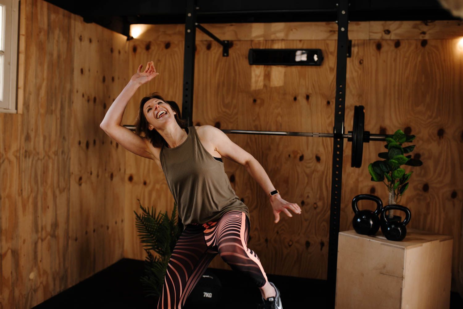 Een persoon in een tanktop en een legging met print lacht terwijl ze zich uitrekt in een sportschool met houten muren en fitnessapparatuur.