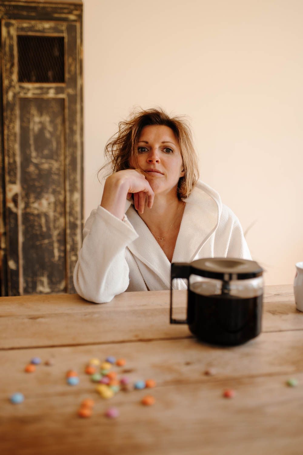 Een persoon in een wit gewaad laat zijn kin op zijn hand rusten aan een houten tafel met koffie en verspreide snoepjes.