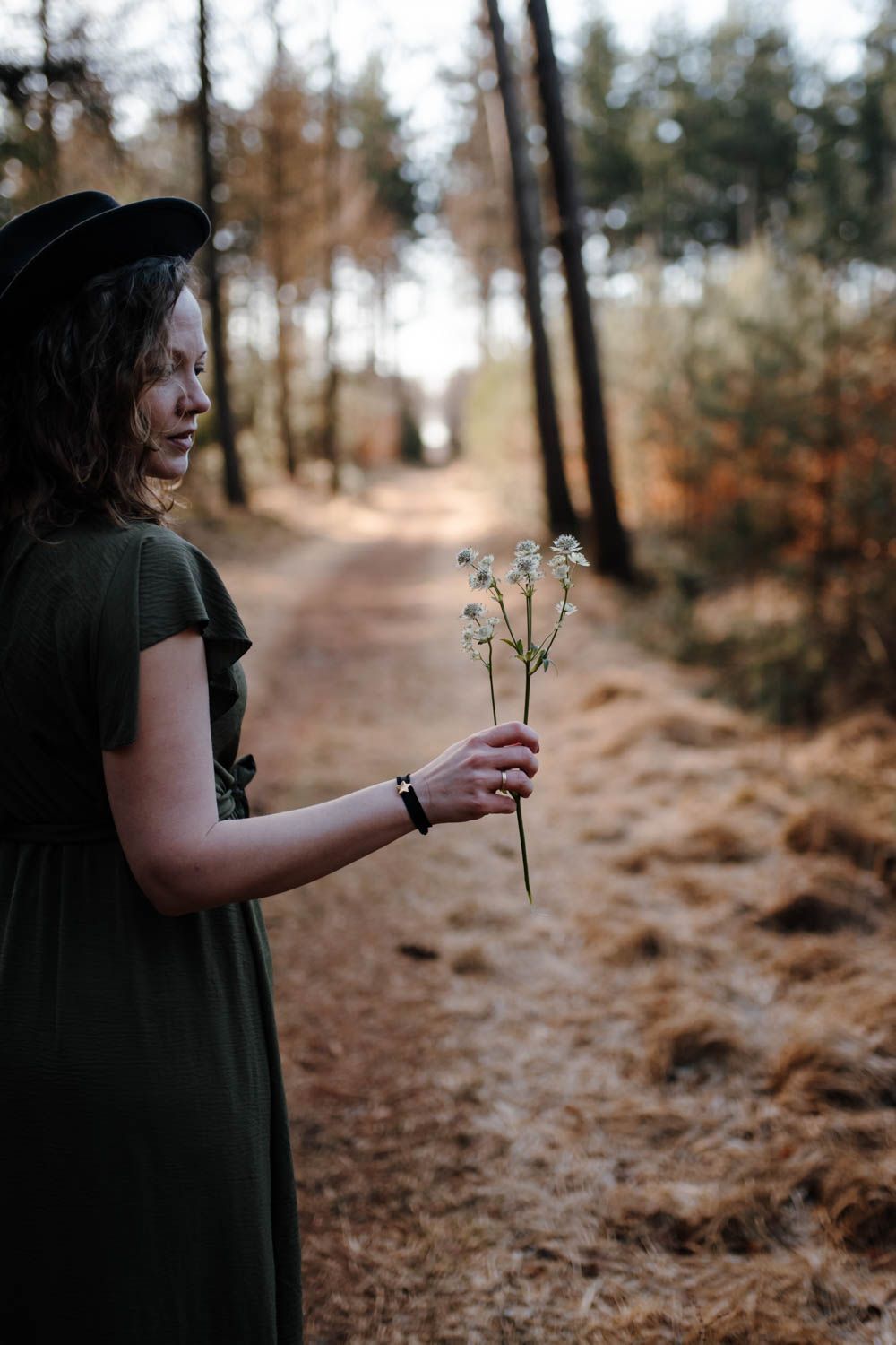 Een persoon in een donkergroene jurk en hoed staat in een bos en houdt drie kleine, gedroogde bloemen in zijn hand.